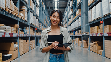 An asian woman smiling is holding a tablet and stylet in a warehouse