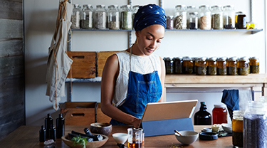 A woman working on her laptop computer while standing in an organic products workspace surrounded by jars of ingredients.